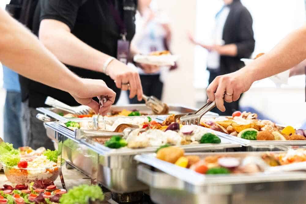 People serving themselves at a buffet line with a variety of dishes, including salads and hot entrees, using tongs to pick up food from chafing dishes