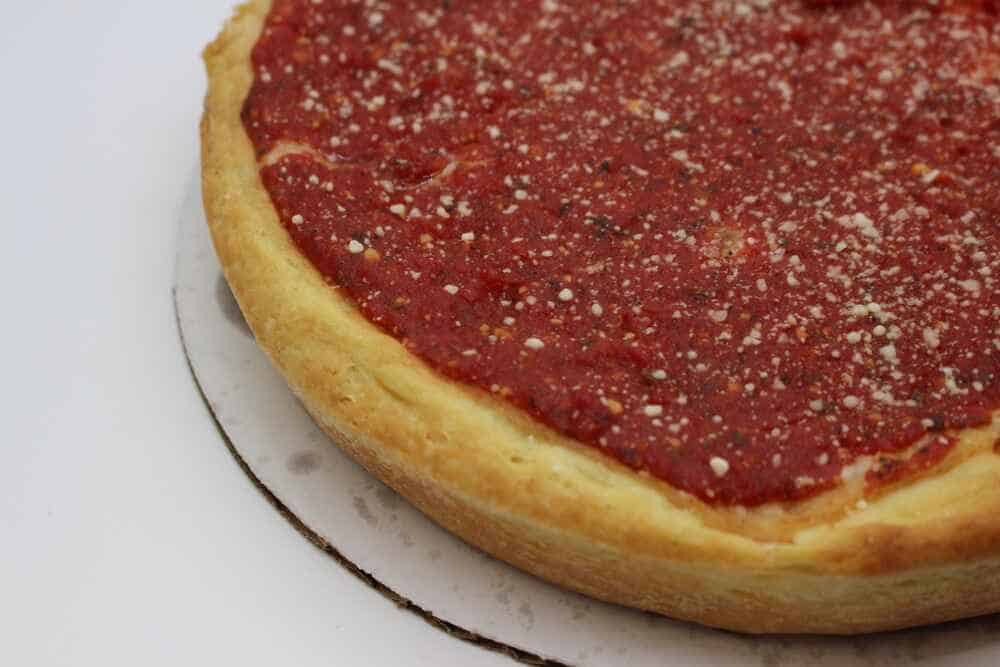 Chicago Deep Dish Delight Close-up of a Chicago-style deep-dish pizza with a high golden crust and a thick layer of vibrant red tomato sauce, sprinkled with grated Parmesan cheese, presented on a white surface.