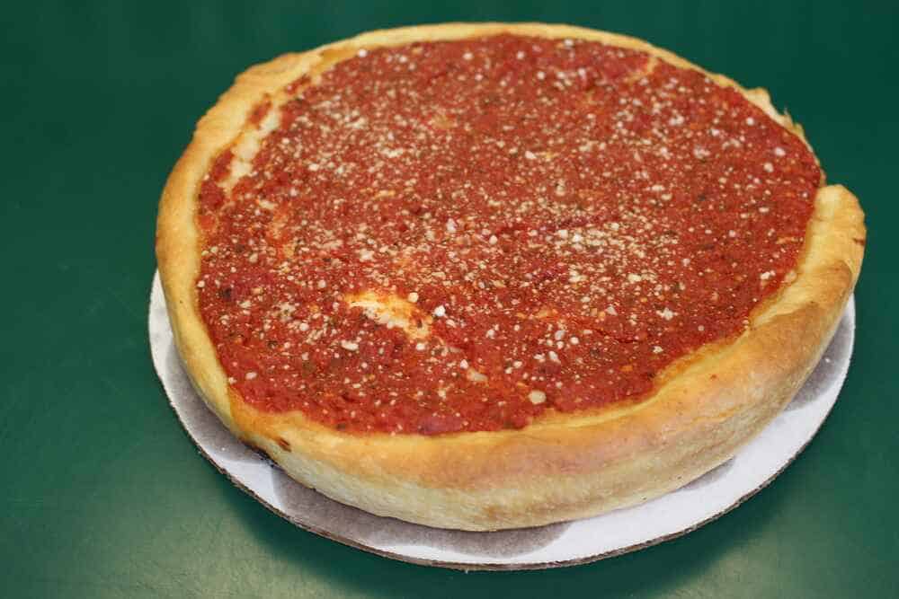 Classic Chicago-style deep-dish pizza on a green table, showcasing a thick and flaky crust with a generous layer of chunky tomato sauce and grated Parmesan cheese on top, served on a cardboard pizza tray.